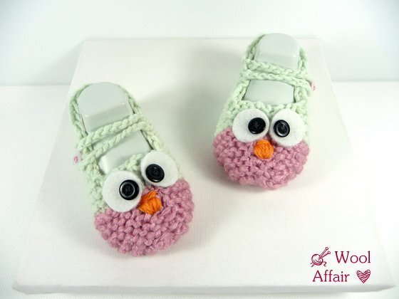 Zwei mintgrün-rosa gestrickte Babyschuhe mit Eulenaugen und orangefarbenem Schnabel auf weißem Hintergrund