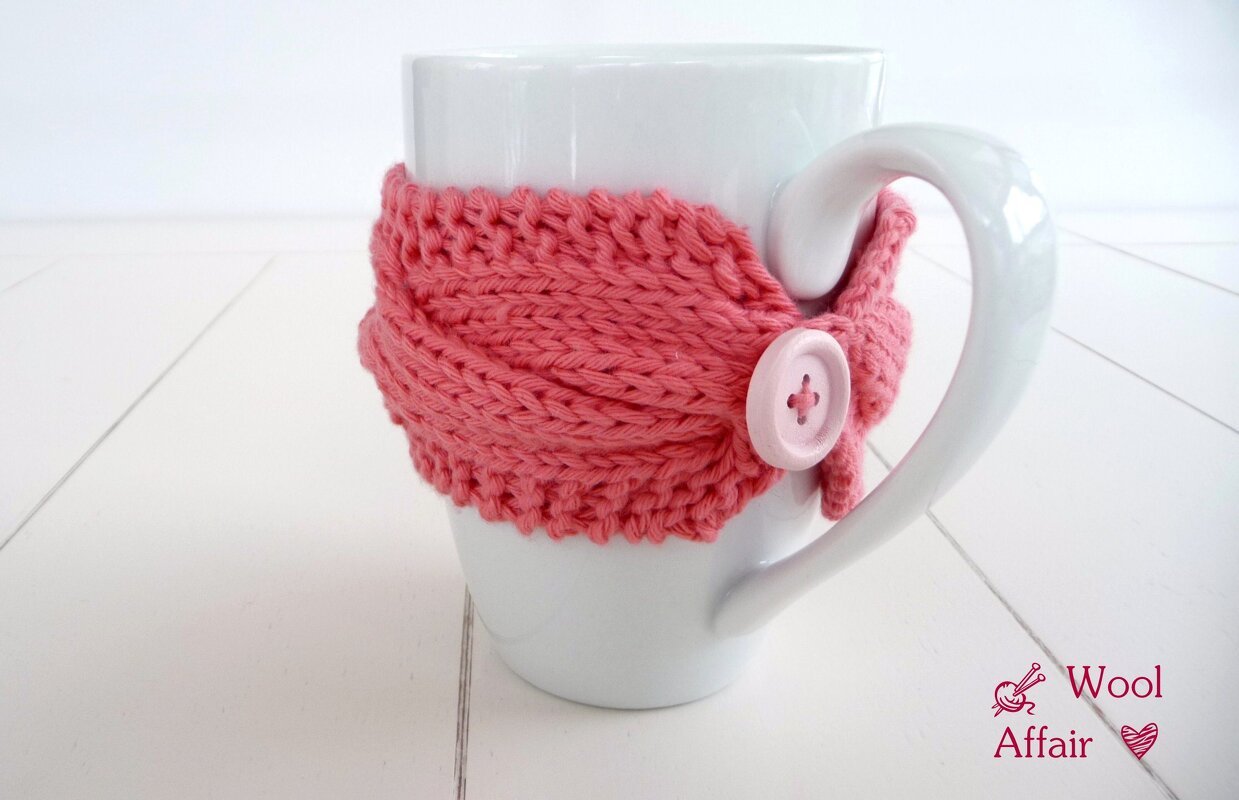 Weiße Tasse mit lachsrosa gestricktem Tassenwärmer mit Zopfmuster und Knopf