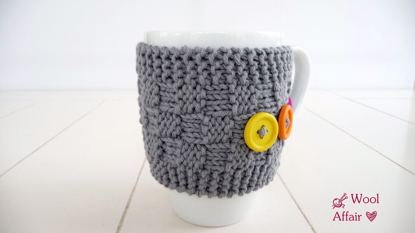 Weiße Tasse mit grauer, gestrickter Hülle mit Zopfmuster und drei bunten Knöpfen