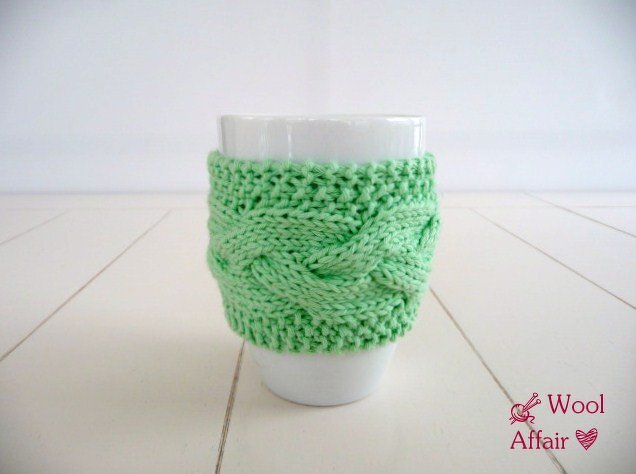 Weiße Tasse mit hellgrüner gestrickter Tassenhülle mit Zopfmuster auf hellem Hintergrund.
