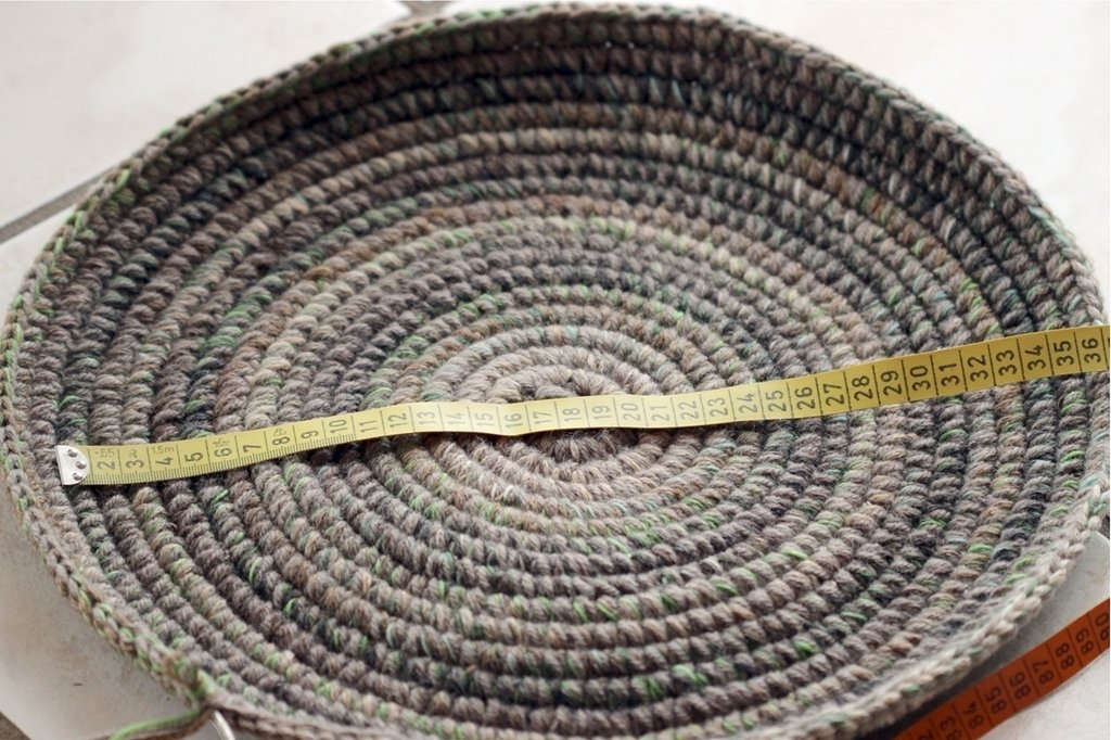 Top-down view of a round crocheted basket in grey variegated yarn with a yellow measuring tape across the center