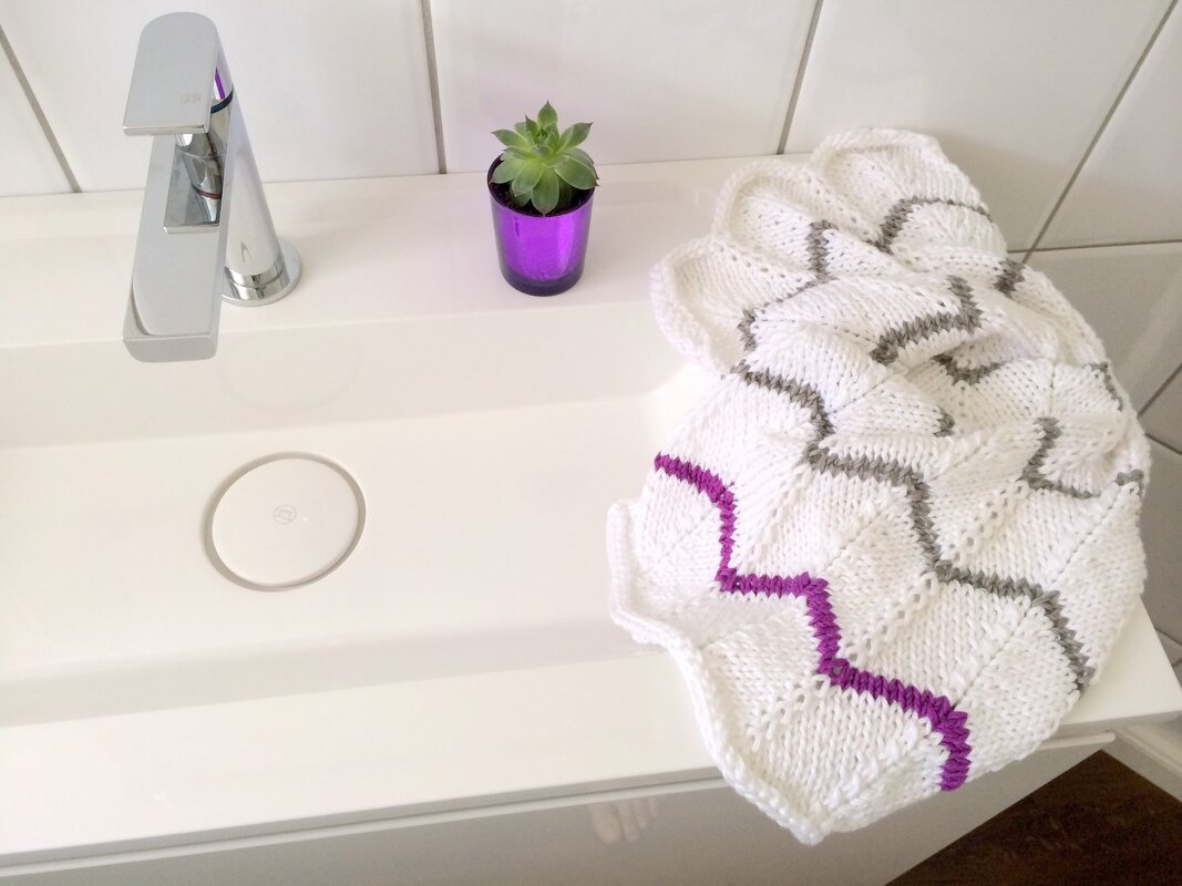 Weißes gestricktes Tuch mit grauen und violetten Zickzackstreifen auf dem Rand eines Waschbeckens neben kleinem Sukkulenten-Topf
