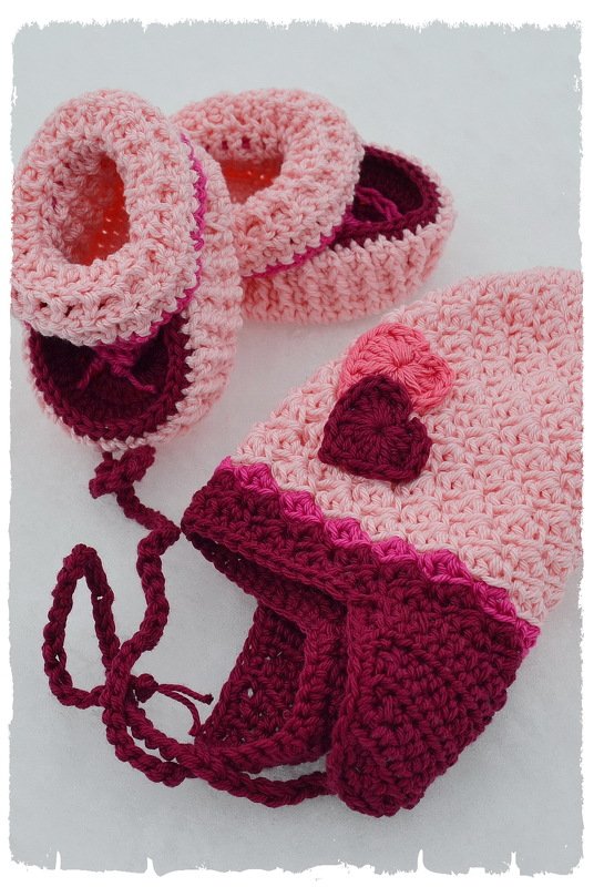 Gehäkeltes Babyset mit rosa Mütze mit Ohrenklappen und Bindebändern sowie passenden rosa-weinroten Schühchen mit Herzapplikationen.