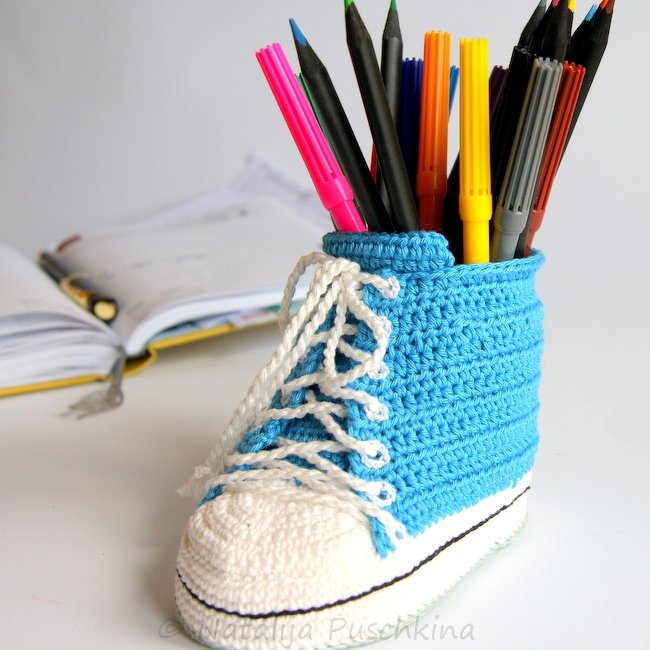 Gehäkeltes blau‑weißes Utensilo in Sneaker‑Form, gefüllt mit bunten Stiften, auf hellem Schreibtisch vor geöffnetem Notizbuch.