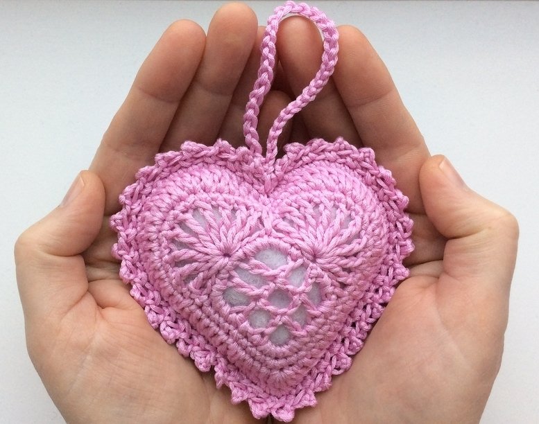 Hände, die ein rosafarbenes gehäkeltes Herz mit Aufhängeschlaufe halten