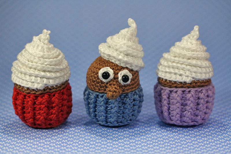 Drei gehäkelte Mini-Cupcakes mit weißer Cremehaube; der mittlere zeigt ein Eulen-Gesicht, die Unterteile sind gerippt in Rot, Blau und Lila.