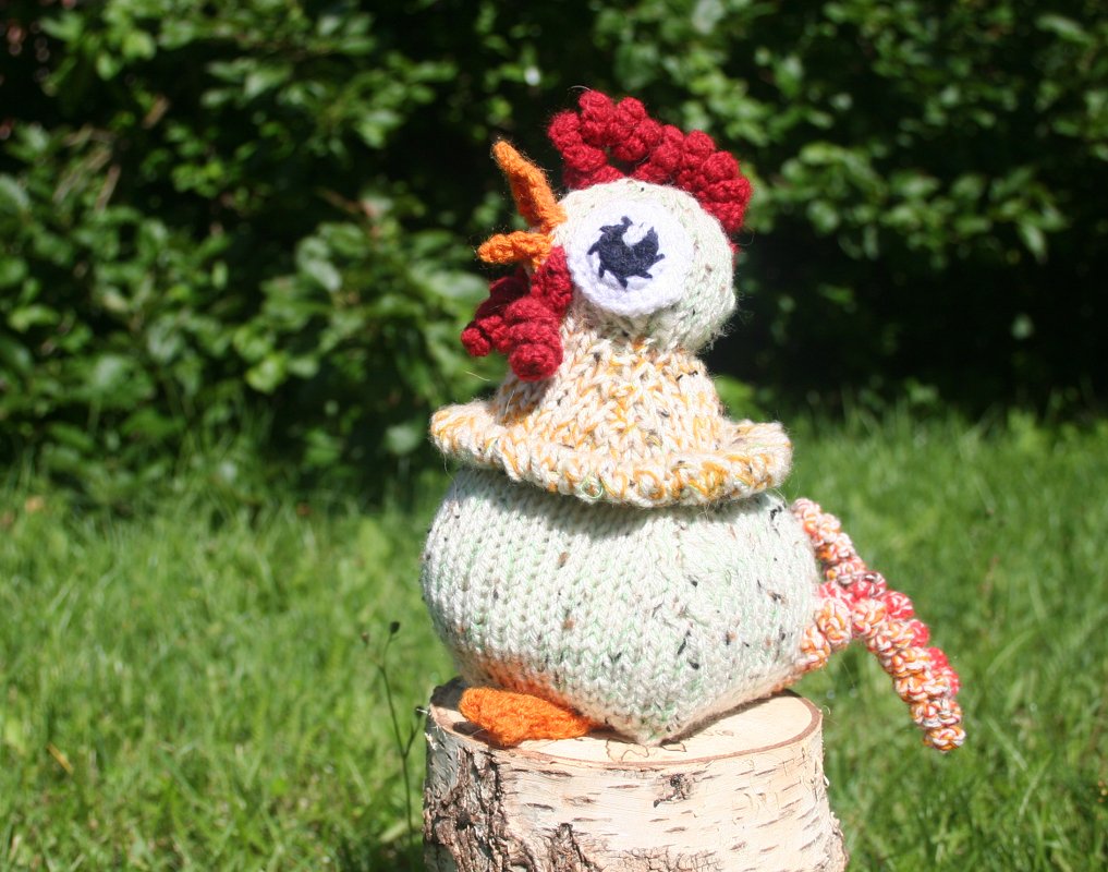 Hellgrünes gestricktes Amigurumi-Huhn mit rotem Kamm und orangefarbenem Schnabel auf Holzstumpf vor grünem Hintergrund.