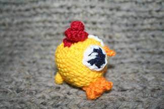 Nahaufnahme eines gelben, gestrickten Amigurumi-Kükens mit rotem Kamm und orangefarbenen Füßen auf grauer Oberfläche.