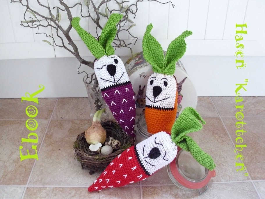 Drei gehäkelte Amigurumi‑Karotten mit Hasengesichtern in Rot, Lila und Orange mit grünen Blättern
