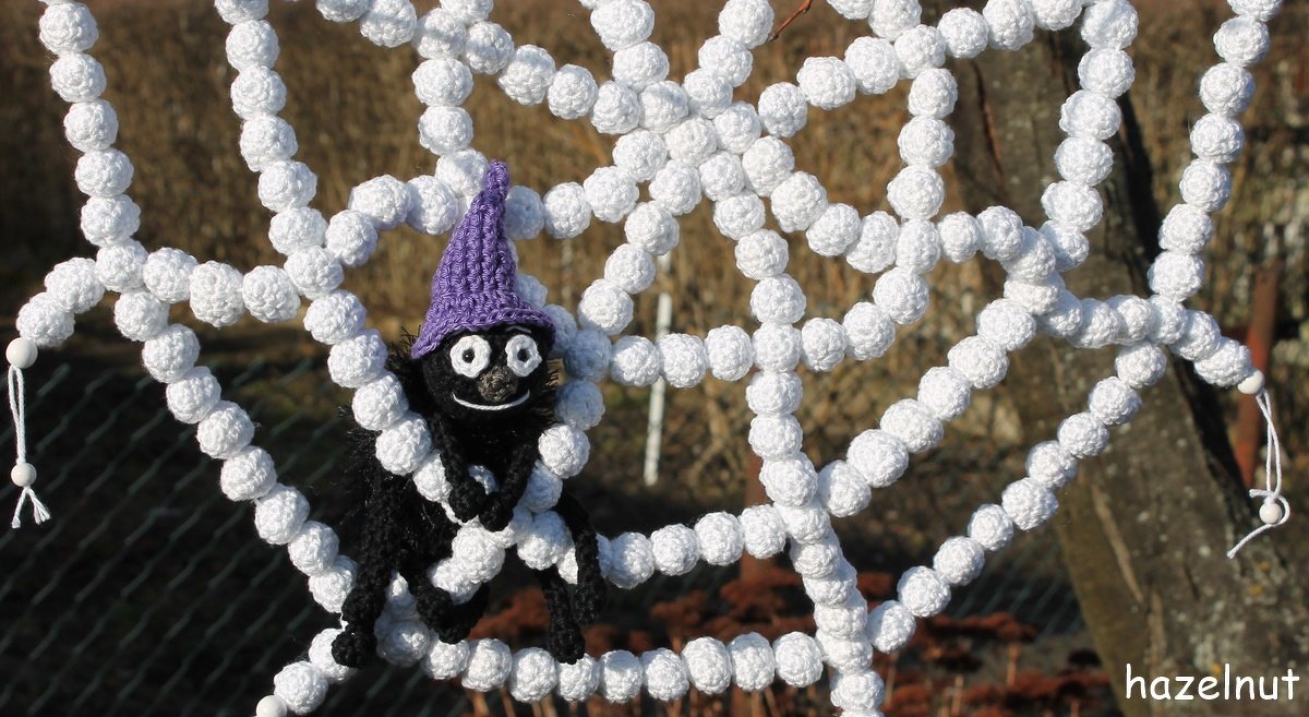 Schwarze gehäkelte Spinne mit lila Hexenhut auf weißem gehäkeltem Spinnennetz