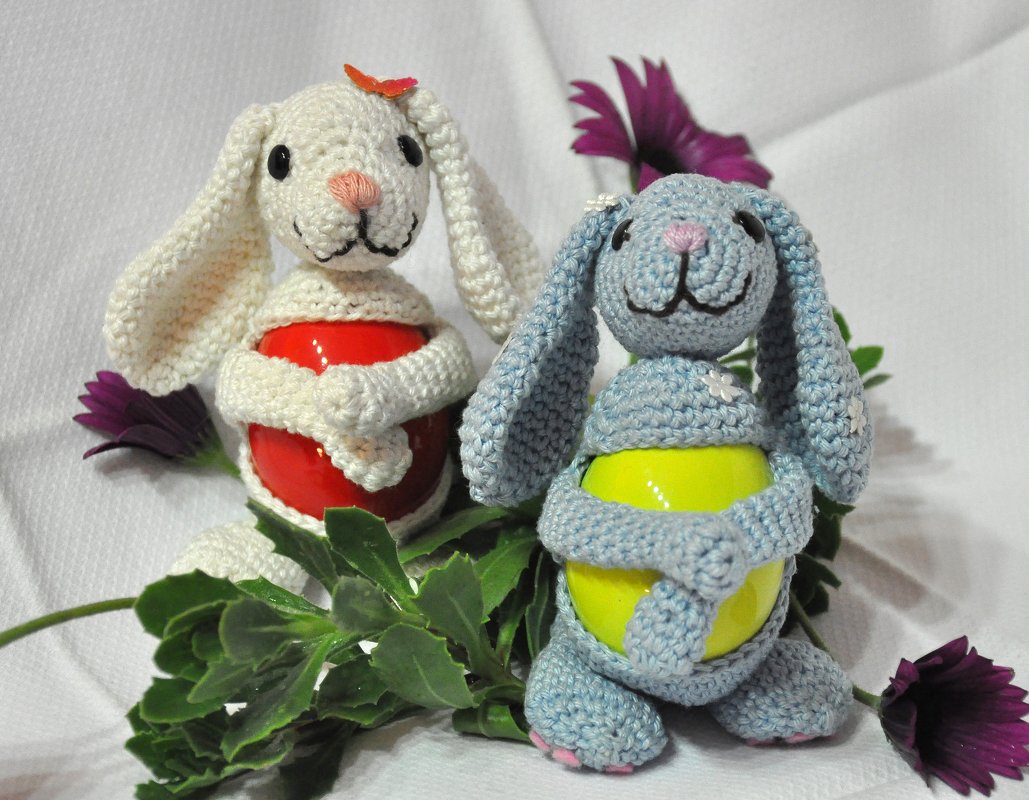 Zwei gehäkelte Osterhasen-Eierwärmer, ein cremefarbener mit rotem Ei, ein hellblauer mit gelbem Ei, dekoriert mit Blättern und Blumen.