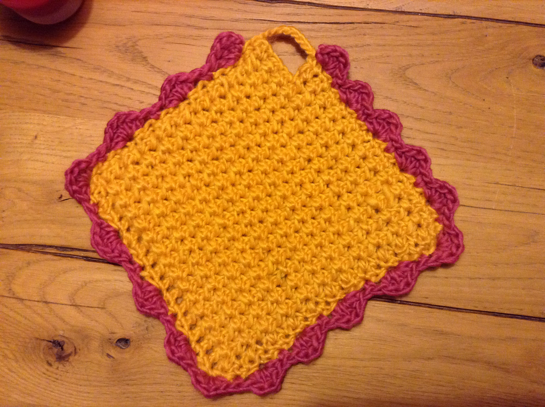 Quadratischer gelber Topflappen mit pinkfarbener Muschelkante und Aufhängeschlaufe auf Holz.