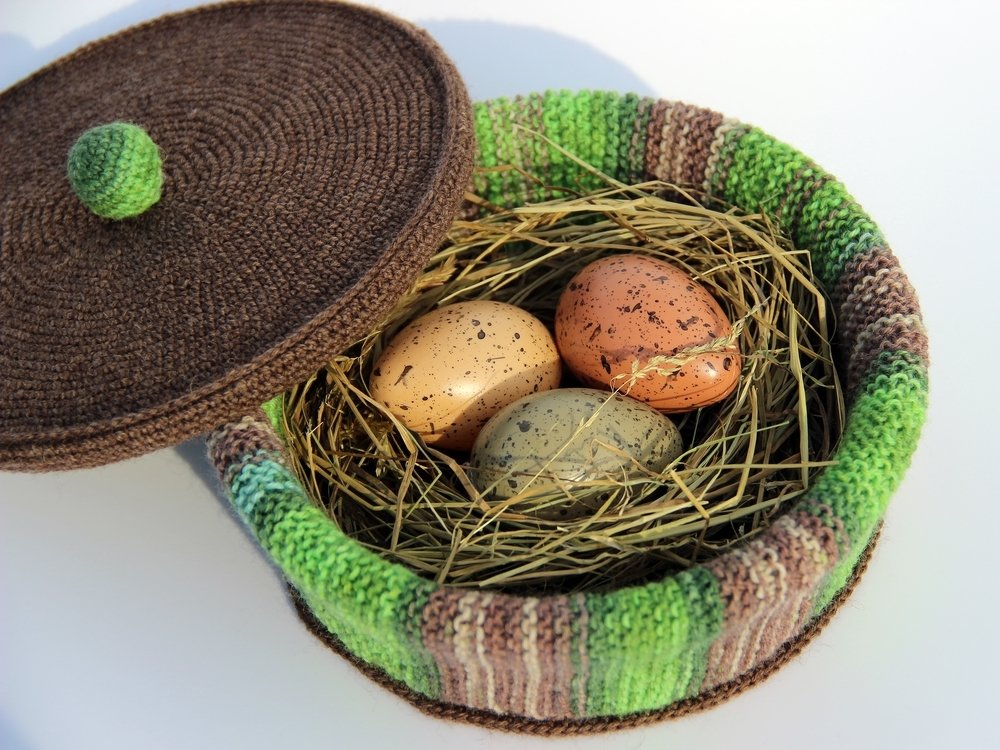 Rundes gehäkeltes Utensilo mit grün-braun gestreiftem Rand und braunem Deckel; innen Stroh und drei Dekoeier