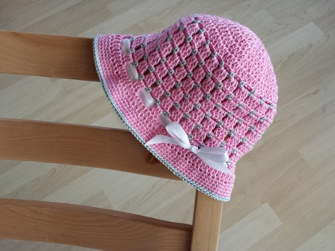 Rosa gehäkelte Babymütze mit weißer Schleife auf Holzstuhl