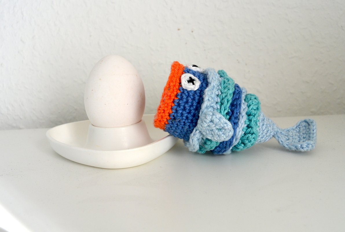 weißes Ei im Eierbecher neben gehäkeltem blauen Fisch‑Eierwärmer mit orangefarbenem Rand und Knopfaugen