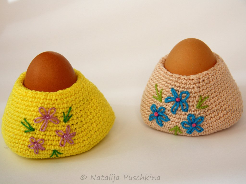 Zwei pastellfarbene gehäkelte Eierwärmer mit blauen und rosa Blumenstickereien und braunen Eiern