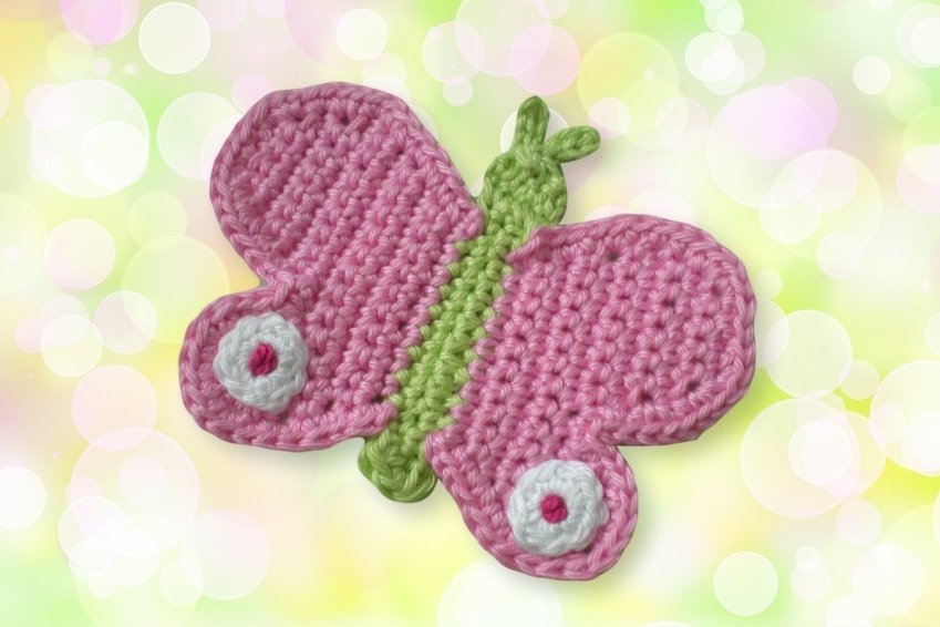 Gehäkelte Schmetterlings-Applikation mit rosa Flügeln, grünem Körper und weißen Kreisdetails vor hellem Bokeh-Hintergrund