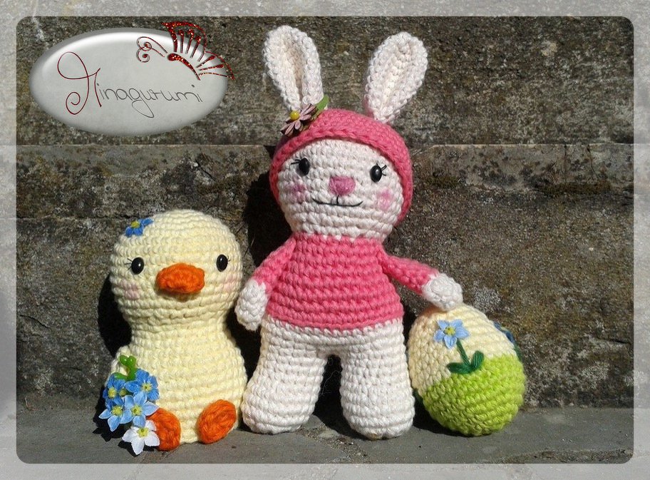 Gehäkelter weiß-rosa Hase mit Mütze neben gelbem Küken mit blauen Blumen vor steinerner Wand