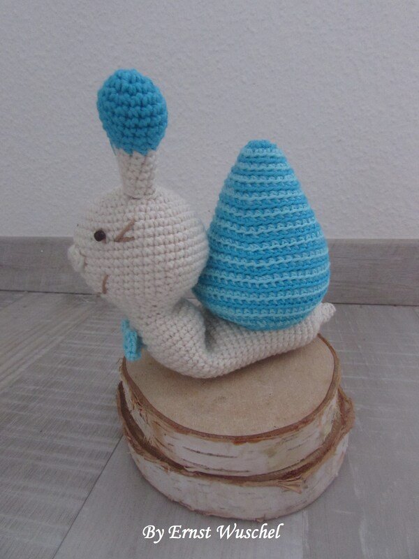 Gehäkelte Amigurumi‑Schnecke creme mit blau gestreiftem Gehäuse auf Holzscheiben