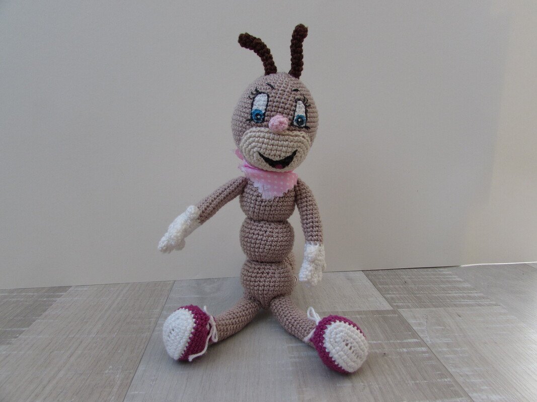Gehäkelte Amigurumi-Raupe mit rosa Halstuch, blauen Augen und magenta-weißen Füßen, sitzend auf Holzfußboden.