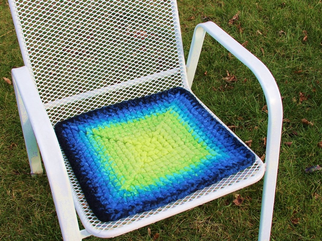 Quadratisches Häkelfilz-Sitzkissen mit grün‑türkis‑blauem Verlauf auf weißem Metallstuhl