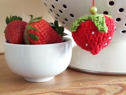 Gehäkelte Erdbeere mit grünen Blättern als Anhänger neben weißer Schale mit frischen Erdbeeren