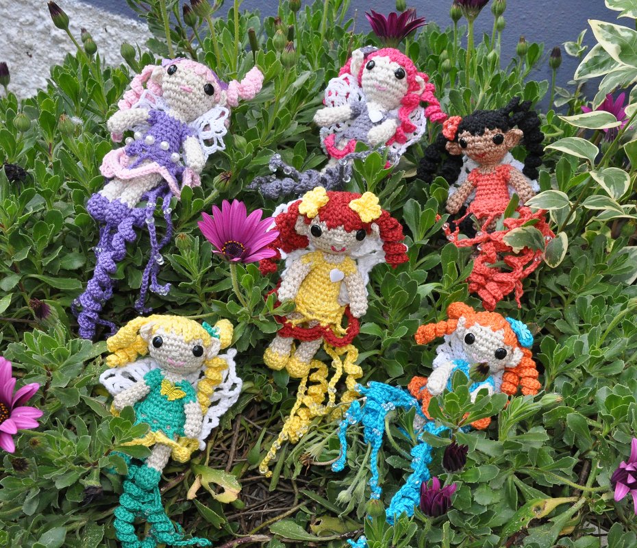 Sieben bunte gehäkelte Amigurumi-Feen mit Quasten und Flügeln auf grünen Pflanzen verteilt