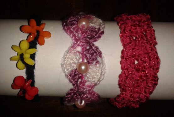 Drei handgefertigte Armbänder: dunkles Band mit bunten Blumen‑Applikationen, rosa‑weißes Häkelarmband mit Perlen, dichtes rotes Häkelarmband.