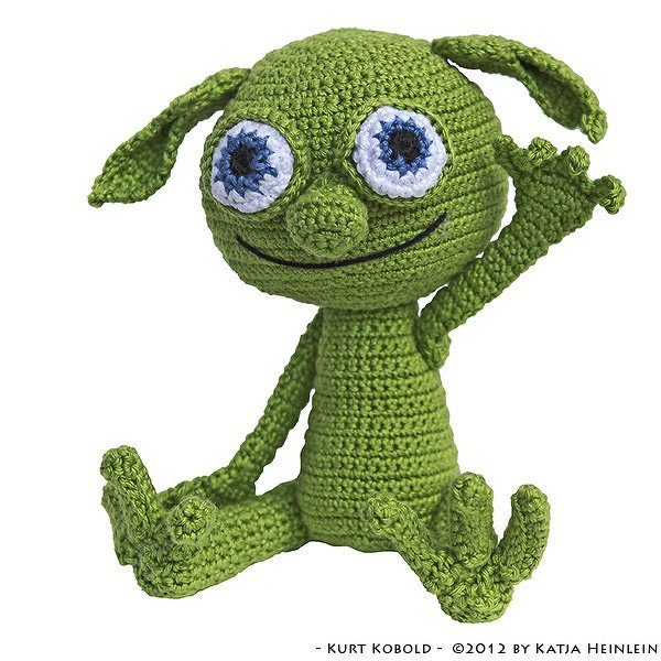 Green crocheted amigurumi creature sitting with one arm raised, large blue-and-white eyes and embroidered smile