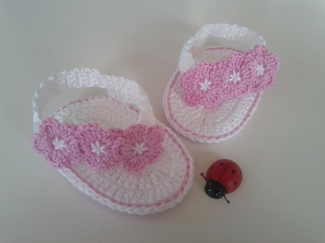 Zwei weiße, gehäkelte Babysandalen mit rosa Blütenapplikationen und einer kleinen Marienkäfer
Figur.