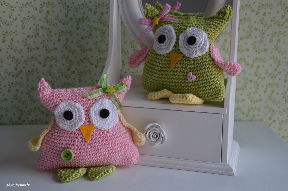 Zwei gehäkelte Amigurumi-Eulen (rosa und grün) mit großen Augen, Schleifen und Knopfapplikationen auf weißer Kommode vor gemustertem Hintergrund.