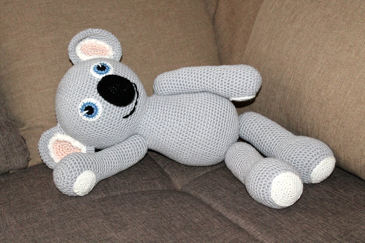 Hellgrauer Amigurumi-Koala liegend mit schwarzer Nase, blauen Augen und weißen Pfotenenden