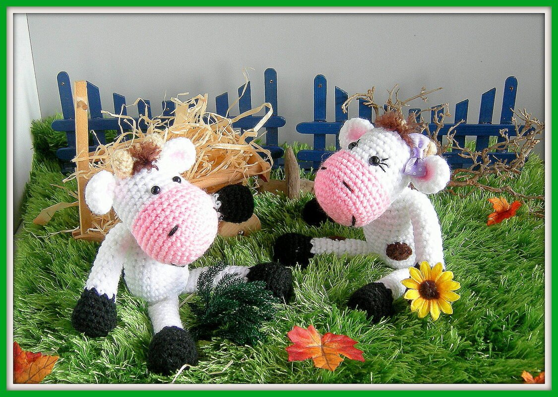 Zwei weiße und schwarze Amigurumi-Kühe mit rosa Schnauzen auf grünem Plüschgras vor blauem Miniaturzaun.