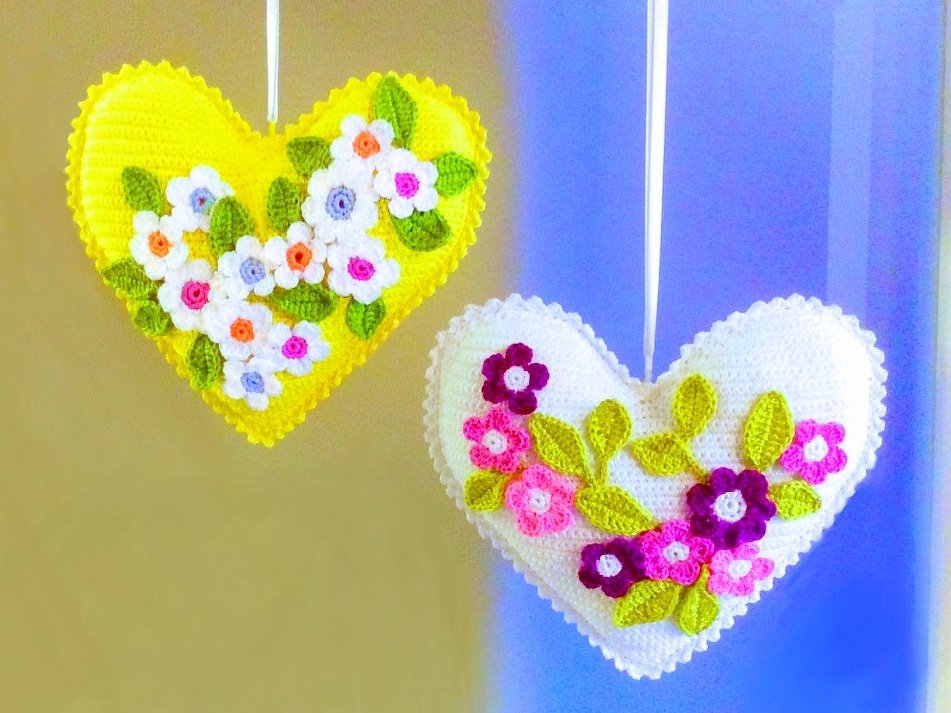 Zwei gehäkelte Herz-Anhänger, gelb und weiß, mit aufgenähten bunten Blumen und grünen Blättern.