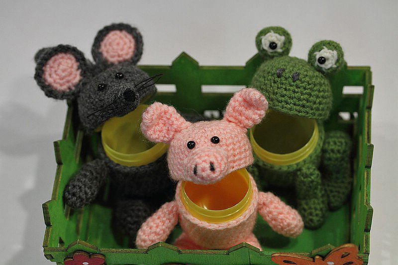 Drei gehäkelte Amigurumi-Tiere (rosa Schwein, graue Maus, grüner Frosch) in einem grünen Holzkasten