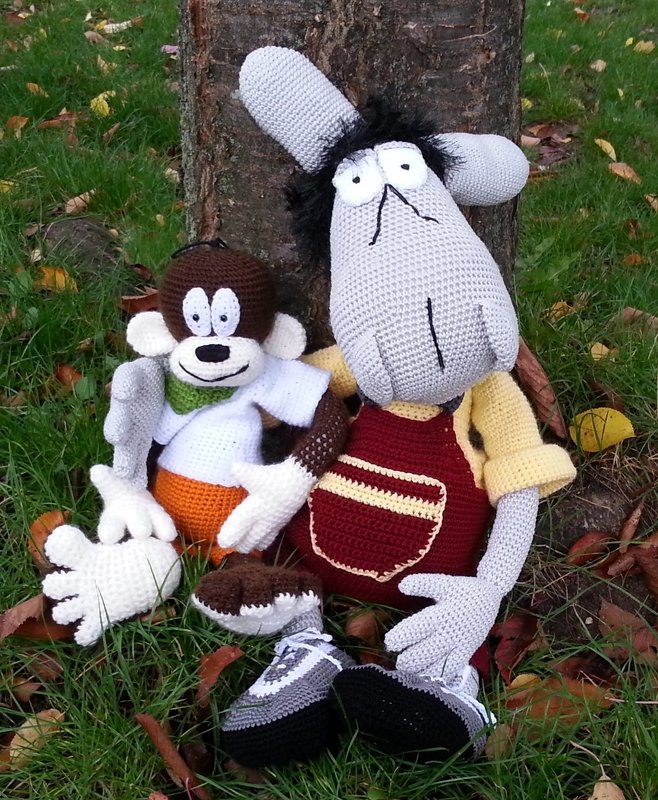 Zwei gehäkelte Amigurumi-Figuren: brauner Affe mit orangefarbener Hose und graue Figur mit Latzhose, sitzend im Gras vor einem Baum