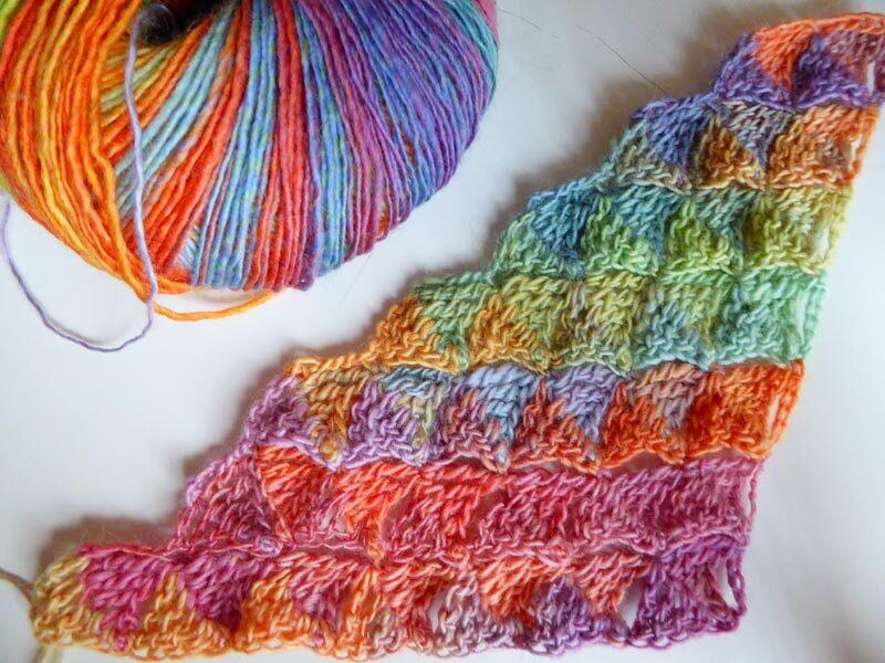 Häkelprobe eines dreieckigen Tuchs mit Muschelmaschen neben einem Knäuel mehrfarbigem Garn