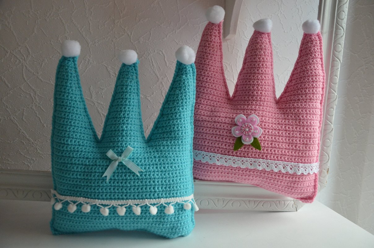 Zwei gehäkelte Kronenkissen in Rosa und Türkis mit weißen Pompons und Zierborten auf hellem Hintergrund.