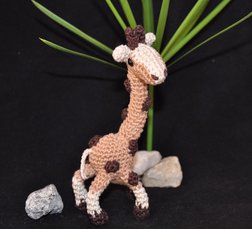 Nahaufnahme einer handgehäkelten Amigurumi-Giraffe in Beige und Braun vor dunklem Hintergrund mit Pflanzenblättern und Steinen.