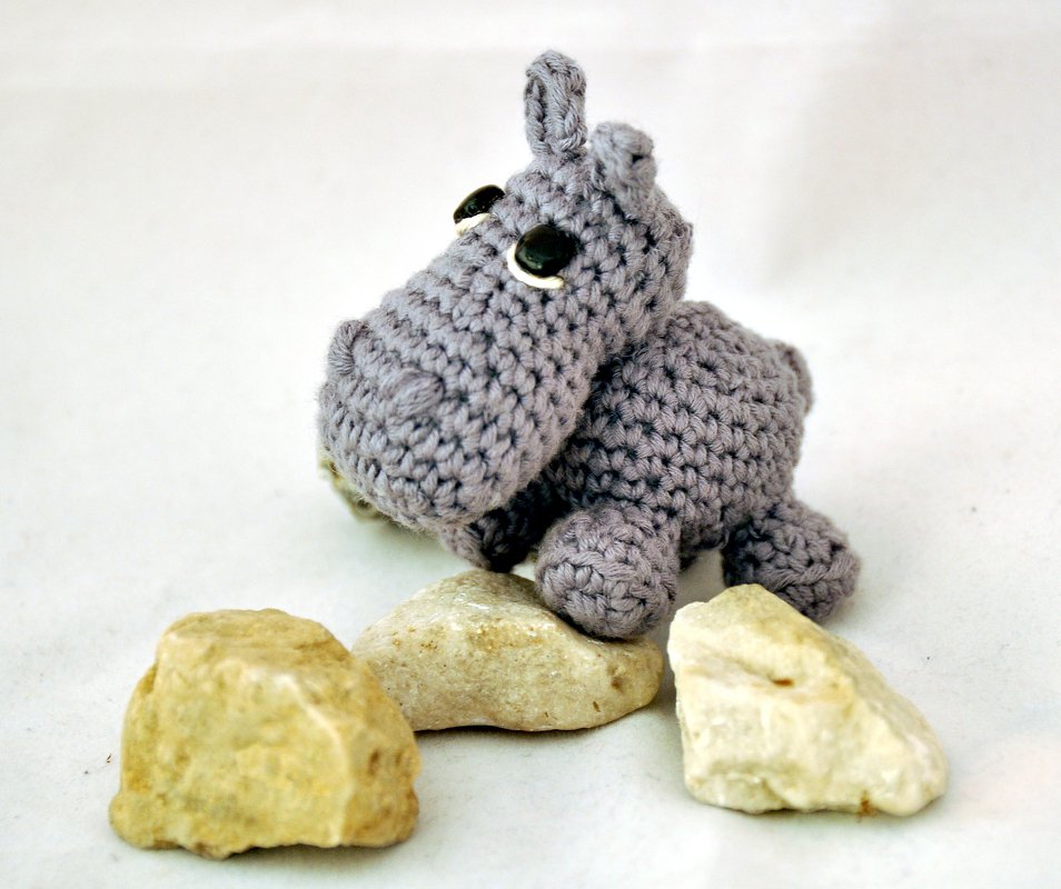 Gehäkeltes graues Amigurumi-Nilpferd mit schwarzen Augen auf hellem Untergrund neben drei Steinen