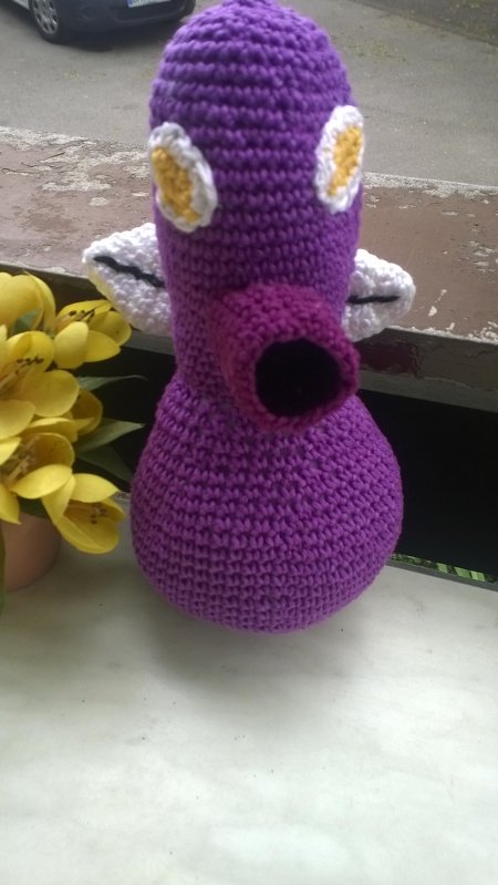 Lila gehäkeltes Amigurumi mit rundem Körper, trichterförmigem Schnabel, weißen Augen und seitlichen Flügelchen