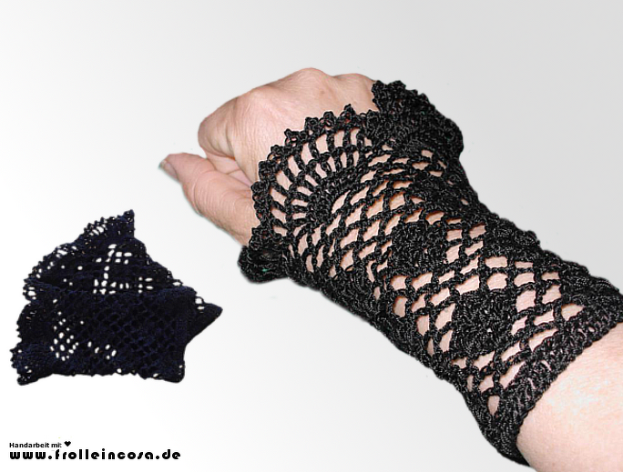 Handgelenk mit gehäkelter schwarzer Armstulpe in filigranem Lochmuster; daneben liegt ein gefaltetes zweites Stück.