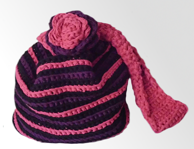 Gehäkelte Tasche mit pink‑lila‑schwarzen Streifen, Blütenapplikation und Trageriemen.