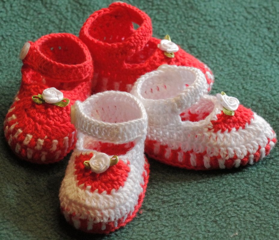 Zwei Paare rot-weißer gehäkelter Babyschuhe (Riemchen und Bootie) mit Rosenapplikationen auf grünem Untergrund.