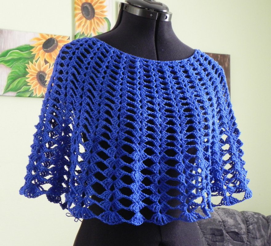 Kobaltblauer gehäkelter Damen-Poncho mit weitem Halsausschnitt und luftigem Lochmuster auf einer schwarzen Schneiderpuppe vor hellgrünem Hintergrund mit Sonnenblumenbildern.