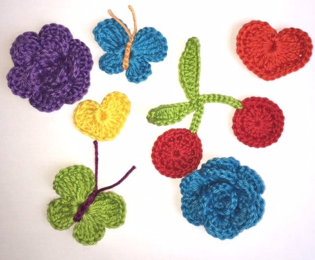 Bunte gehäkelte Applikationen: lila Blume, blauer Schmetterling, rotes Herz, gelbes Herz, grünes Kleeblatt, zwei rote Kirschen und eine blaue Rose.