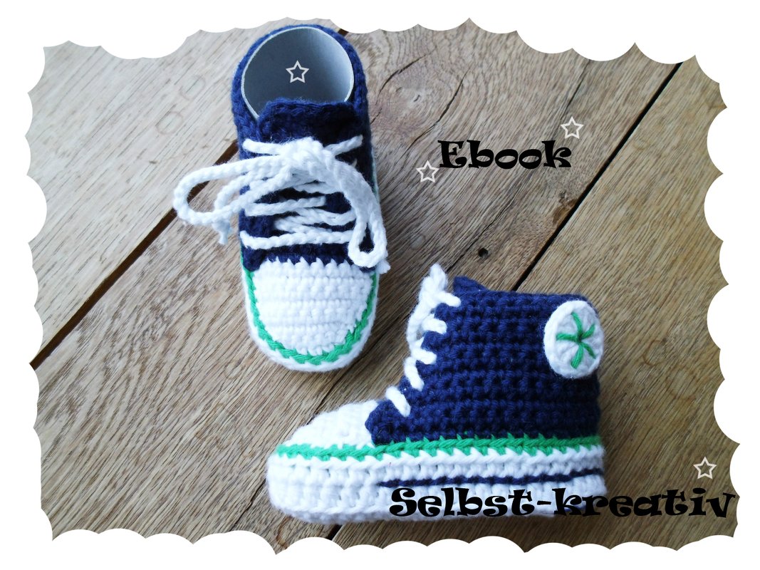 Zwei hohe gehäkelte Babysneaker in Marineblau und Weiß mit grüner Ziernaht auf Holzuntergrund