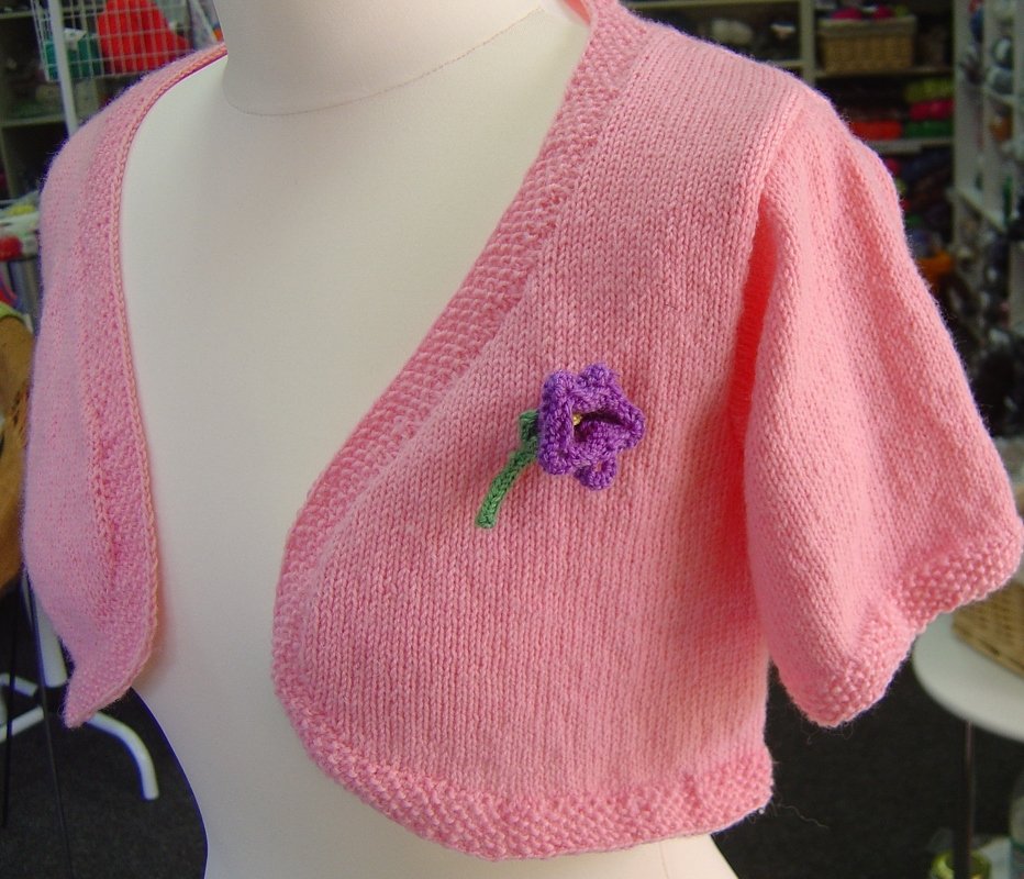 Rosa gestrickter Damenbolero auf einer weißen Schneiderpuppe, verziert mit einer lila gehäkelten Blume.