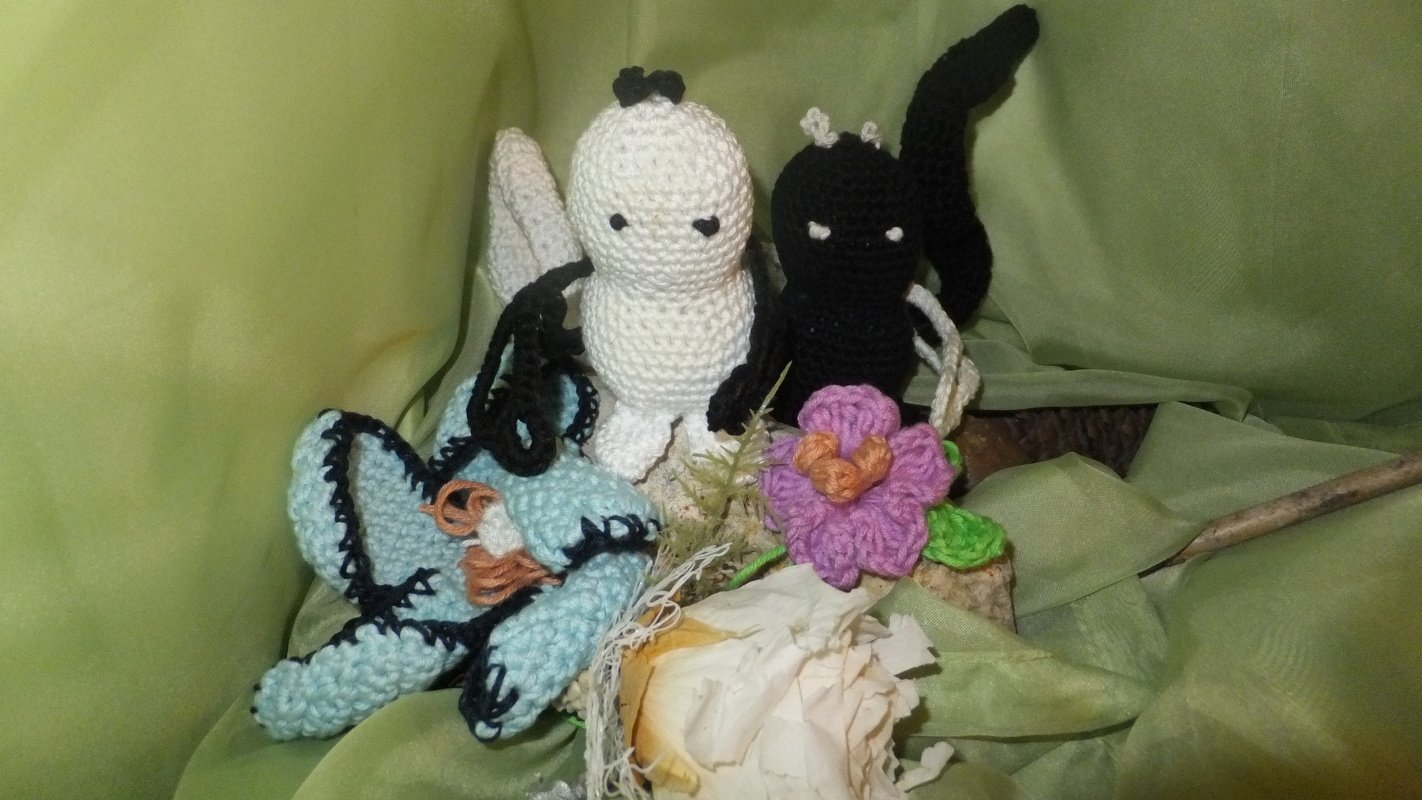 Zwei gehäkelte Amigurumi-Figuren (weiß und schwarz) neben einer lila Häkelblume und einem hellblauen gehäkelten Element auf grünem Stoff.