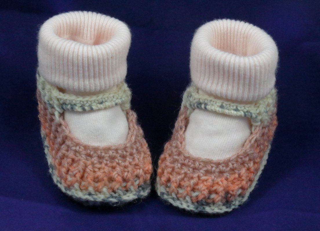 Handgehäkelte Baby-Ballerinas in Pfirsich-, Creme- und Grautönen mit hellen, gerippten Bündchen auf dunkelblauem Hintergrund.